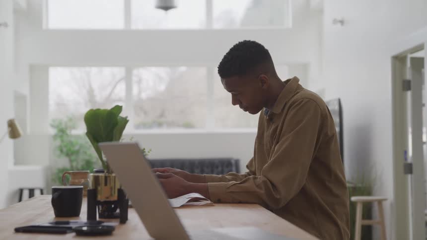 A young man sitting in front of a table reading some invoices and he is wearing a brown jacket