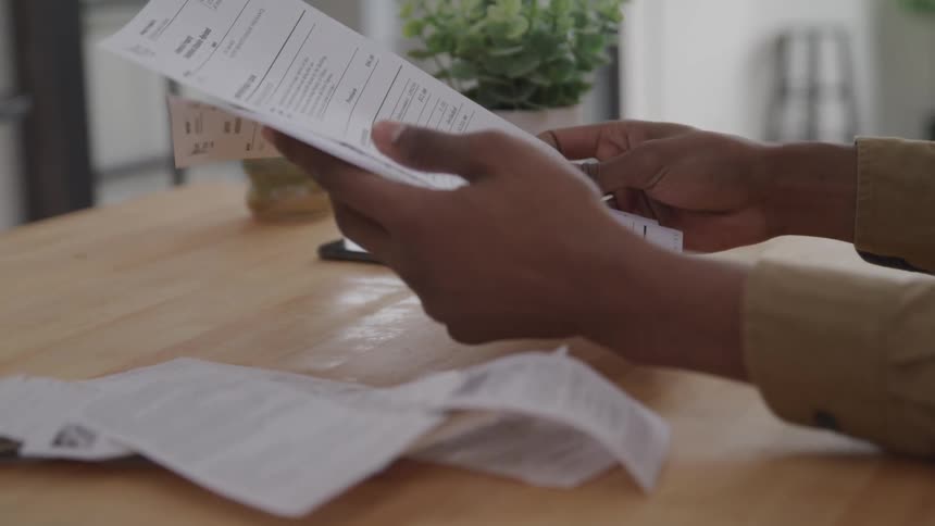 A person is holding and examining several documents at a wooden table with a smartphone and a ceramic cup in the background