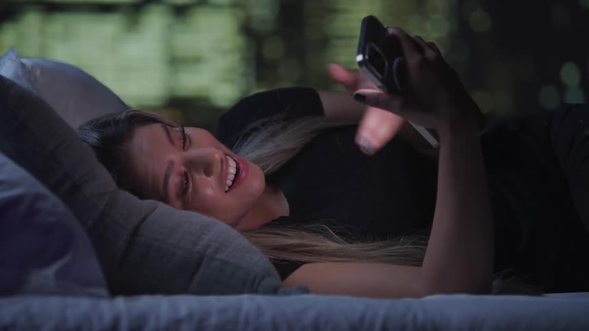 A woman is comfortably lying on a bed propped up on a pillow engaging in a video call on her smartphone
