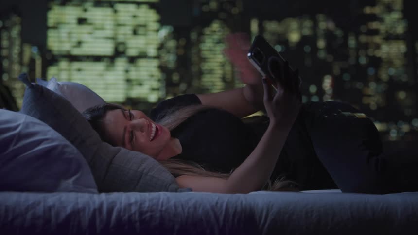 Smiling woman lying in bed making goodbye gesture with hand while on video call