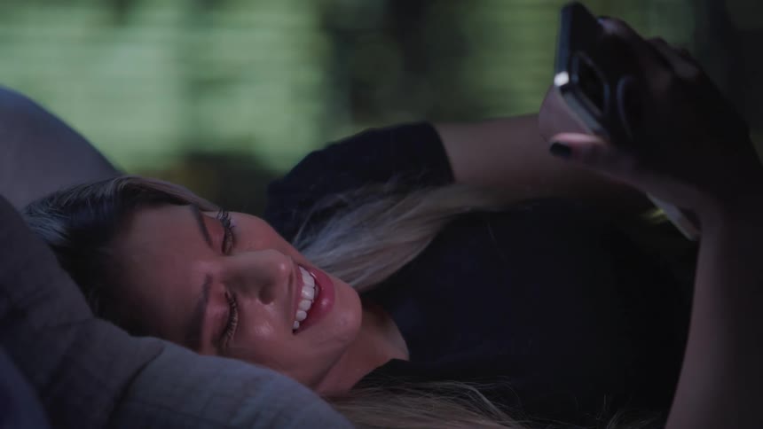 A woman lies comfortably on a bed supported by a pillow while browsing her smartphone