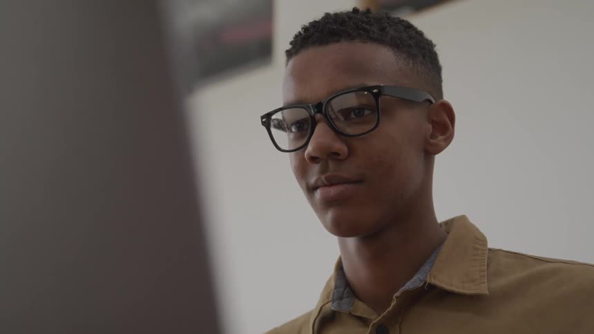 A black teen wearing glasses is smiling while watching the laptop screen
