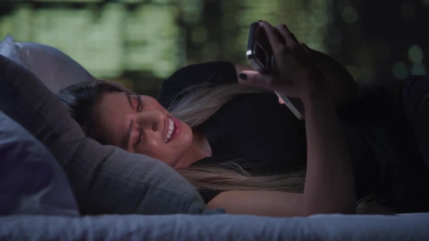A woman lies comfortably on a bed supported by a pillow while browsing her smartphone