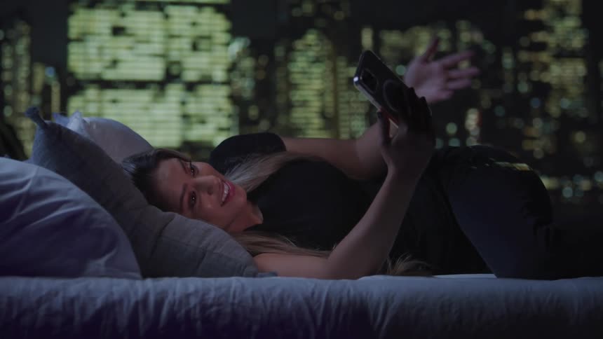 A woman is comfortably lying on a bed propped up on a pillow engaging in a video call on her smartphone