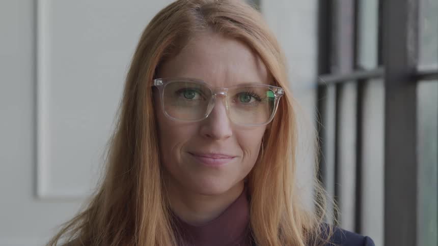 A close up of the face of a woman with long blonde hair wearing glasses near a window