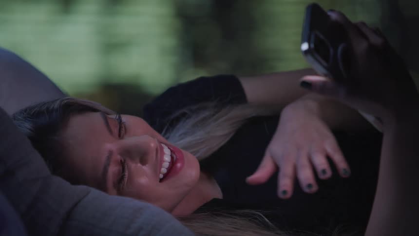 A woman is comfortably lying on a bed propped up on a pillow engaging in a video call on her smartphone
