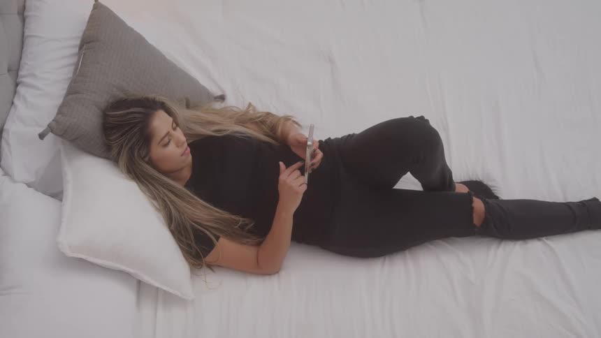 A woman lies comfortably on a bed supported by a pillow while browsing her smartphone