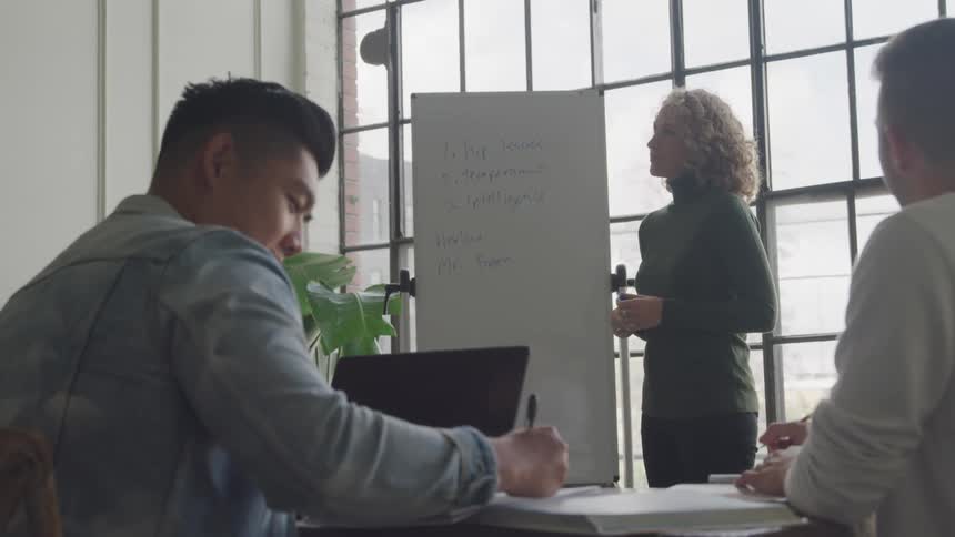 A woman stands in front of a whiteboard with notes while two others sit at a table with a laptop and notebooks engaged in a collaborative session in a room with large windows