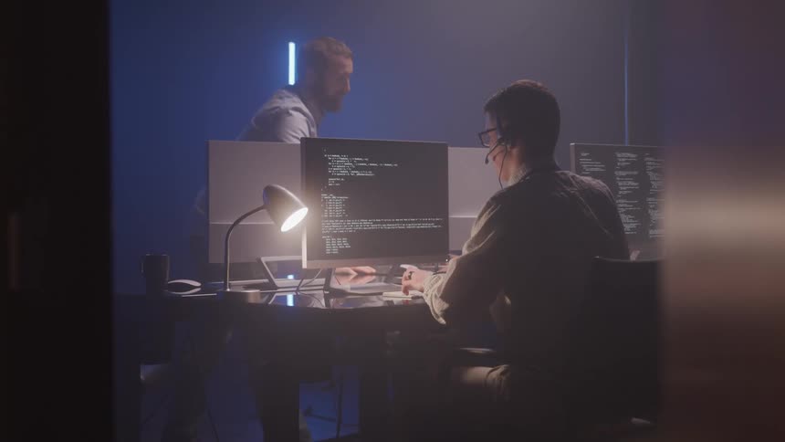Two men work at a desk in a dimly lit room focused on their computers suggesting a call center or tech support setting