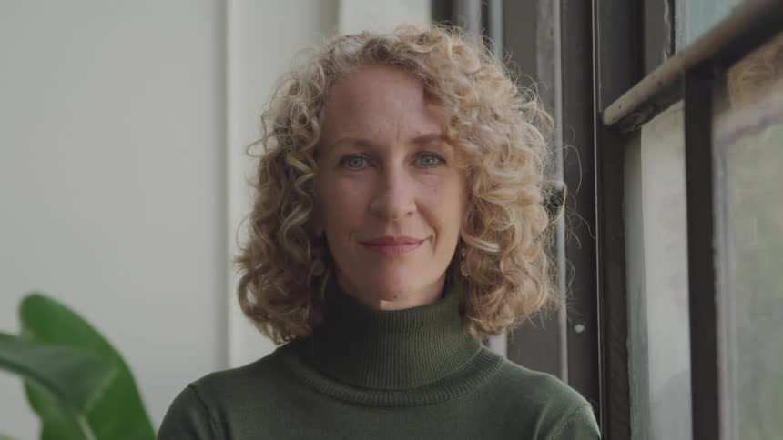 A woman with curly hair and a green sweater with a turtle neck stands next to a window in an office