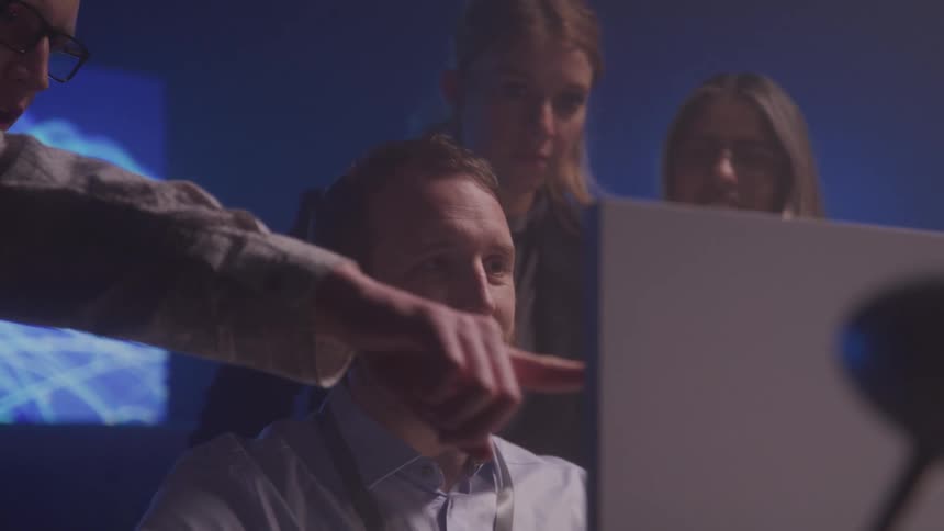 Four individuals in a dimly lit room focus on a coworkers computer collaborating on a task
