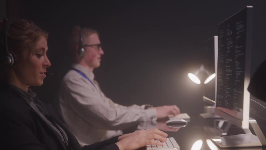 People work at desks in a dimly lit room each focused on their computers while wearing headsets suggesting a call center or tech support setting
