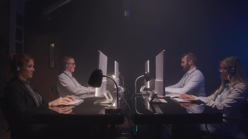 Four individuals work at desks in a dimly lit room each focused on their computers while wearing headsets suggesting a call center or tech support setting