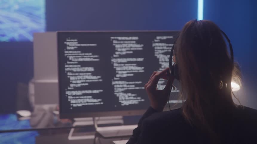 A person wearing a headset sits at a computer displaying lines of code on a dark screen in a modern office