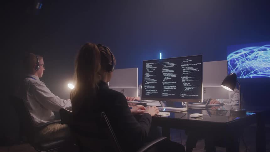 Two people work at a desk in a dimly lit room focused on their computers suggesting a call center or tech support setting
