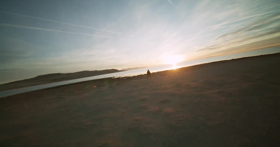 A guy wearing a hat and a red vest is dancing on the sea shore at sunset