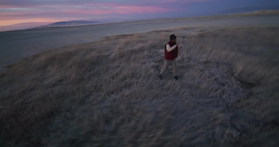 A person dances on a patch of grass near a and a distant coastline under a sky streaked with clouds