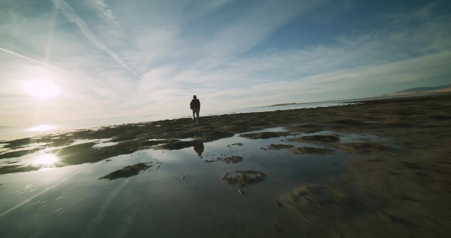 A person dances on a rocky beach nearby and a distant coastline under a sky streaked with clouds