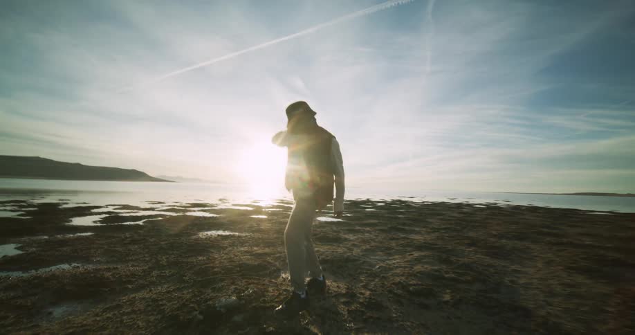 A person dances on a rocky beach with waves crashing nearby and a distant coastline under a sky streaked with clouds