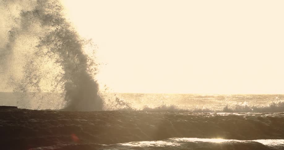A large wave crashes against a rocky shoreline under a bright hazy sky