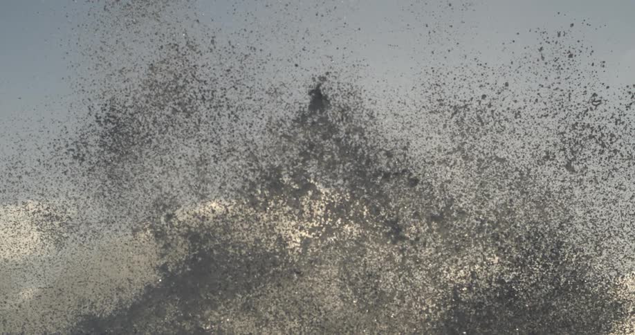 Sea spray bursts against a backdrop of a pale sky at sunset