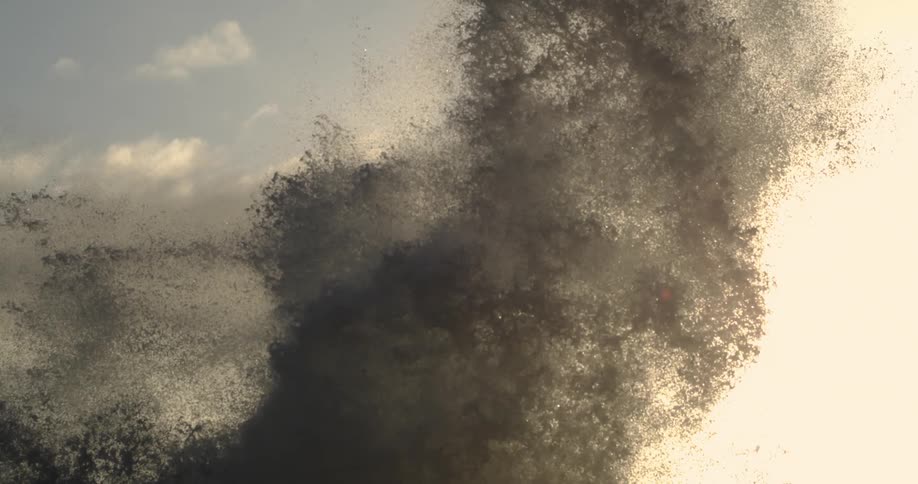 Sea spray bursts from a wave crashing on the coast set against the backdrop of a pale sunset sky