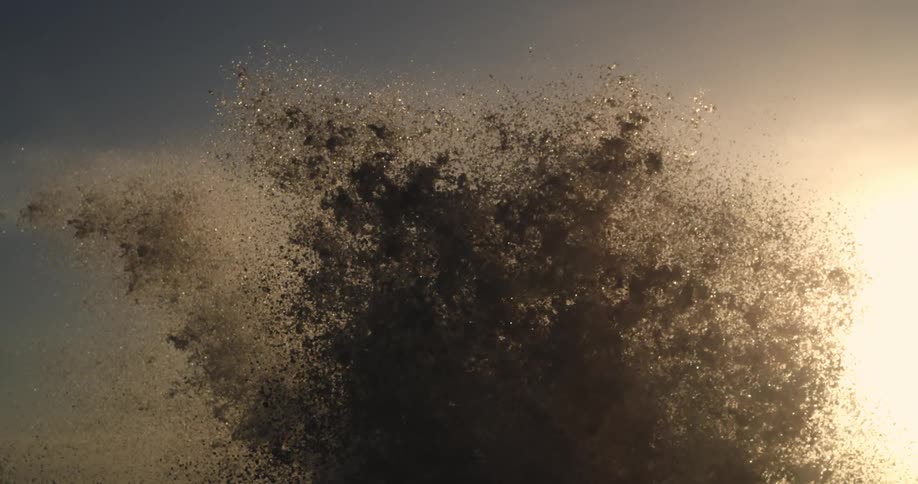 Sea spray bursts against a backdrop of a pale sky at sunset