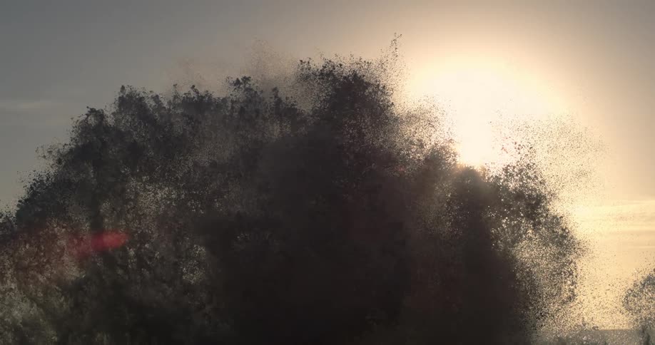 Sea spray bursts against a backdrop of a pale sky at sunset