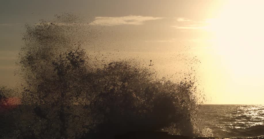 Sea spray bursts against a backdrop of a pale sky at sunset