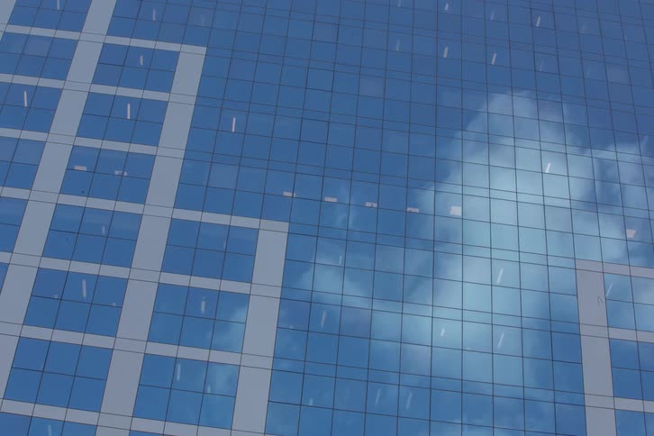A lowangle shot shows a modern buildings facade composed of reflective glass panels mirroring a cloudy sky