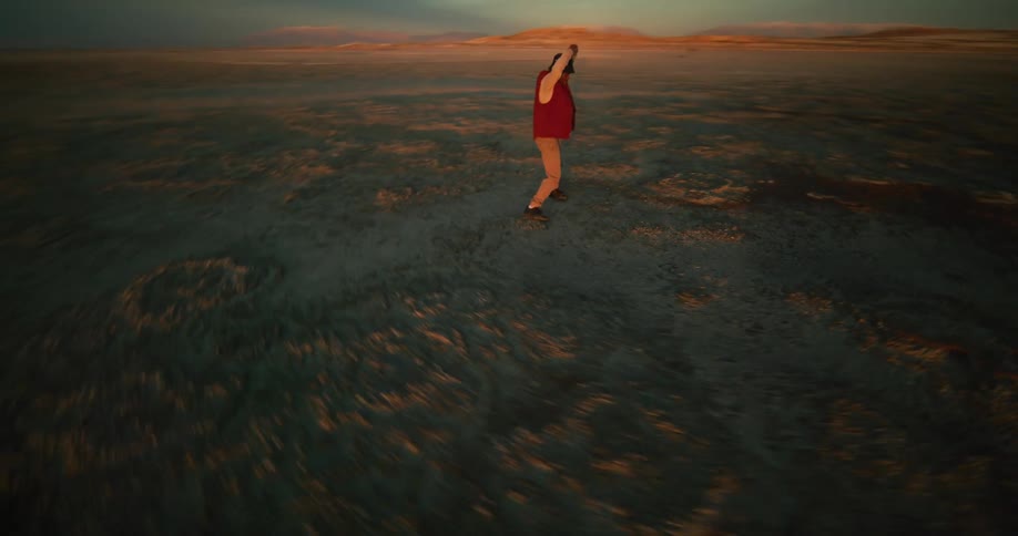 A person dances on a rocky beach nearby and a distant coastline under a sky streaked with clouds