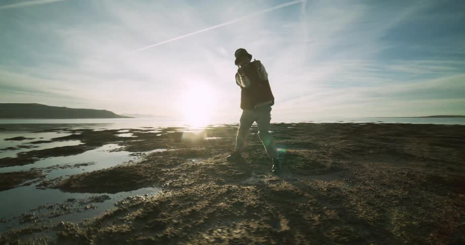 A person dances on a rocky beach nearby and a distant coastline under a sky streaked with clouds