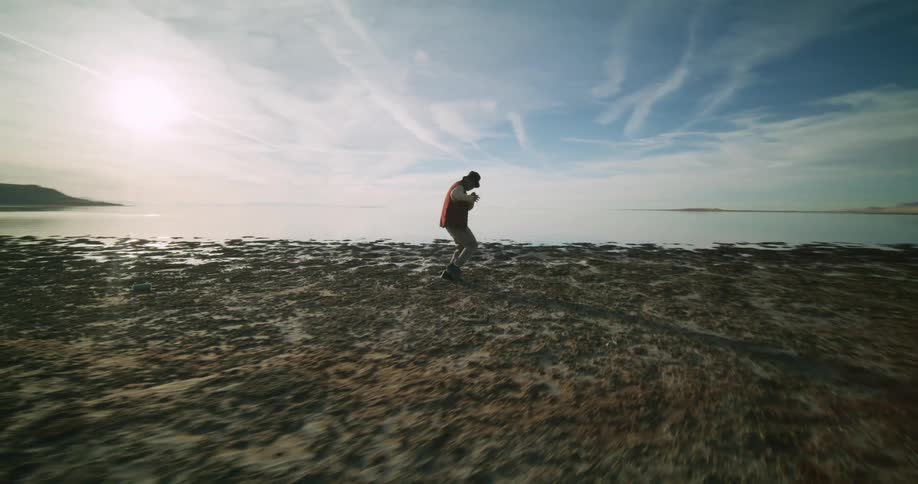 A person dances on a rocky beach nearby and a distant coastline under a sky streaked with clouds