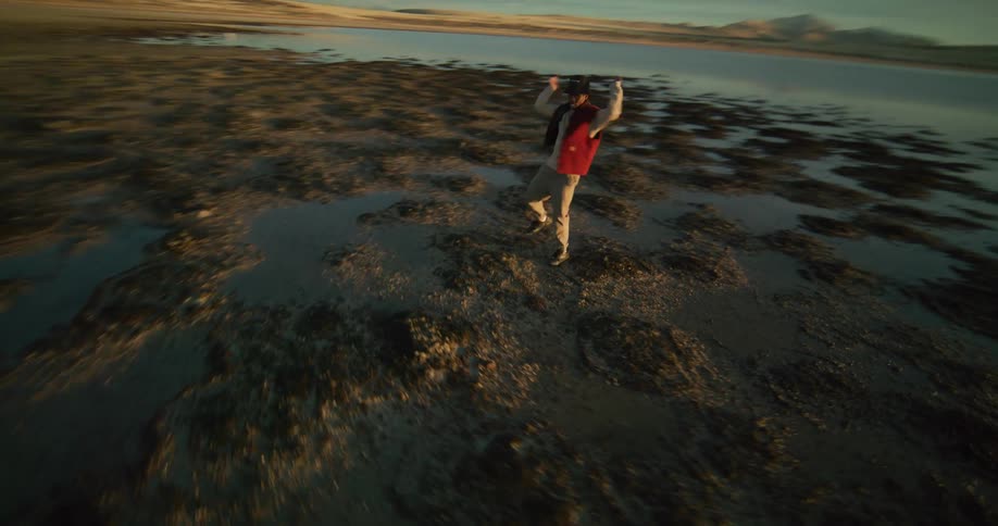 A person dances on a rocky beach nearby and a distant coastline under a sky streaked with clouds