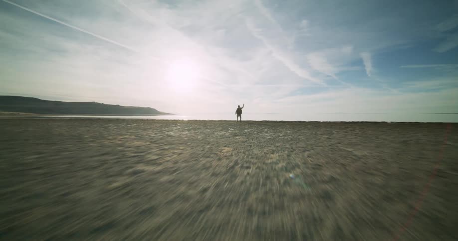 A person dances on a rocky beach nearby and a distant coastline under a sky streaked with clouds