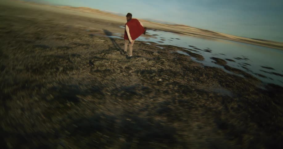 A person dances on a rocky beach nearby and a distant coastline under a sky streaked with clouds