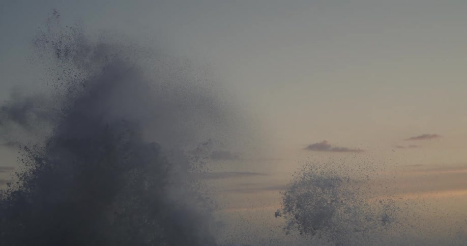 Sea spray bursts against a backdrop of a pale sky at sunset