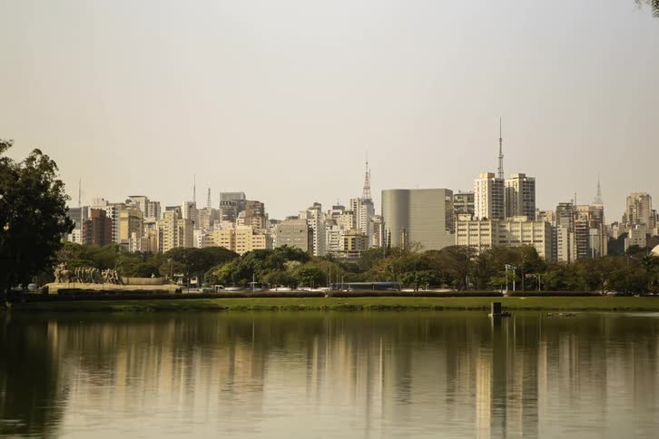 A cityscape skyline rises behind a calm body of water and a line of trees