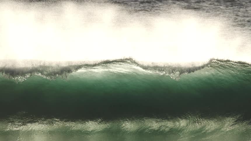 From above a powerful ocean wave curves and peaks as it advances toward the shore beginning to crash and spray white foam as it folds over itself in the sea