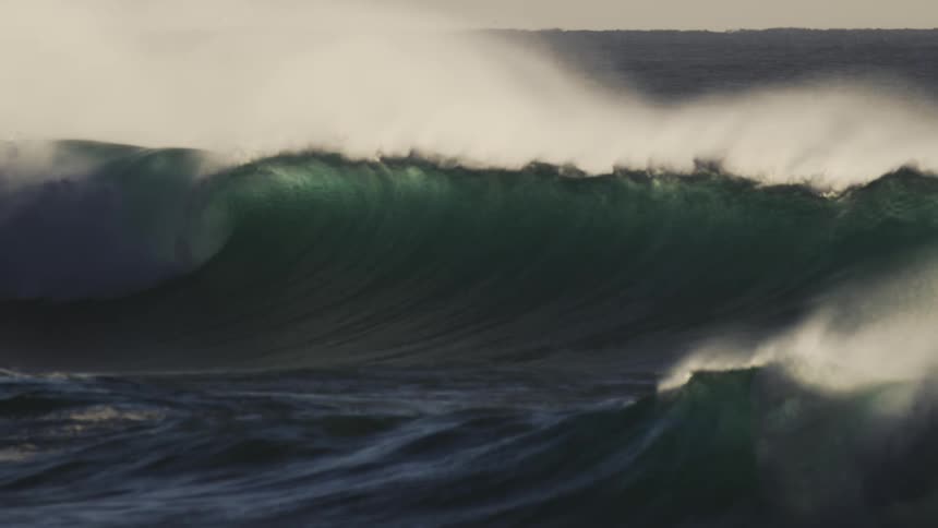 A large dark wave with a white foamy crest towers over the choppy sea