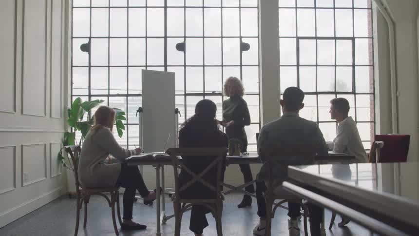 Woman standing and walking around talking to four people sitting around a table
