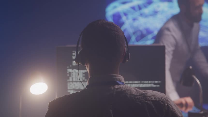 A man stands in front of a man wearing headphones and working on a computer