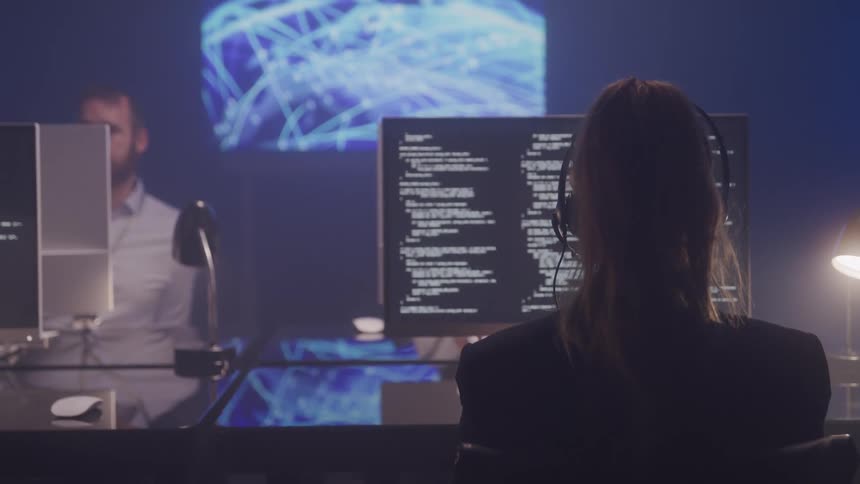 A bearded man stands up from his work desk and taps another coworker on the shoulder while a woman is typing and answering a call