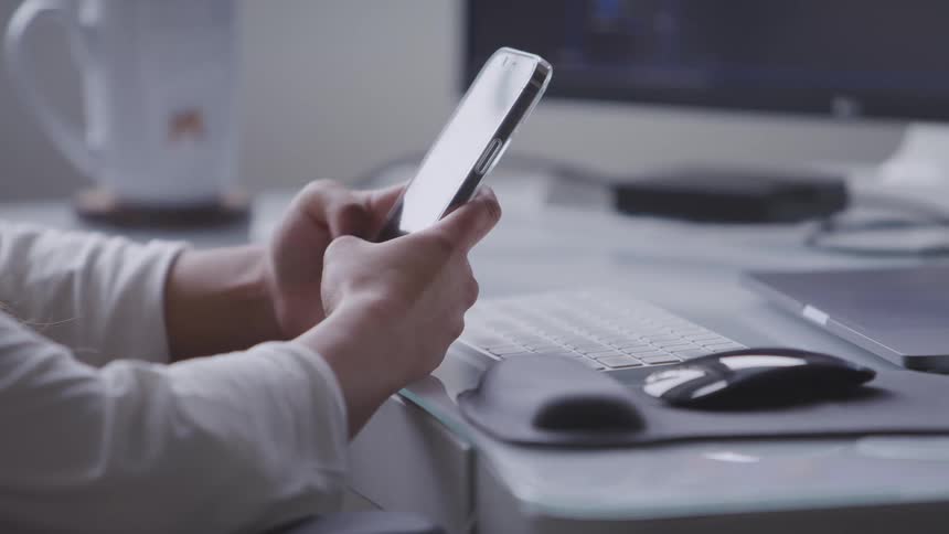 A person texts on a smartphone above a desk with a keyboard mouse and computer peripherals