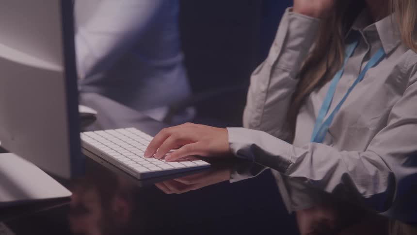 A professional setting where a person wearing a white shirt and a lanyard is typing on a sleek white keyboard with a colleague visible in the background
