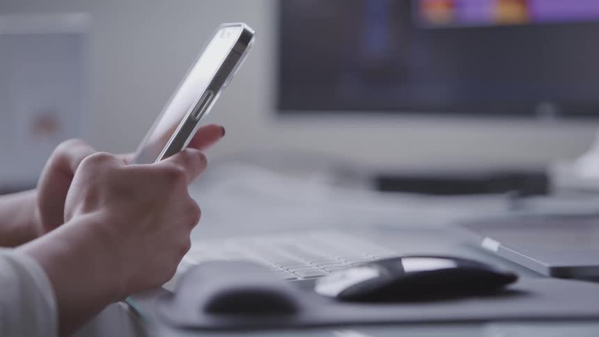 A persons hands texting on a smartphone against a blurred background of a computer screen and desk accessories