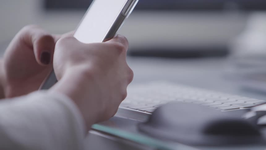 A person texting on a cell phone with a computer keyboard visible in the background