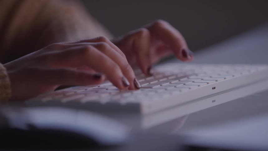 Fingers with dark nail polish type on a white keyboard in a dimly lit setting