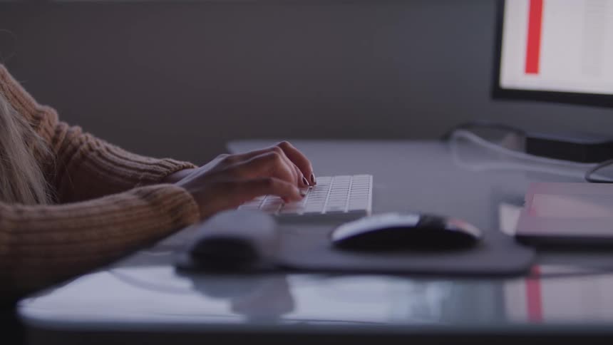 A persons hands type on a keyboard in front of a computer screen with a mouse and mousepad visible on the desk