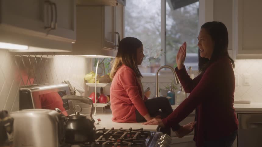 A woman is standing smiling giving high five to a little girl who is sitting in front of her